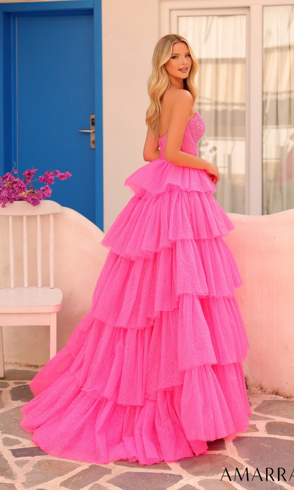 A woman in the Long Formal Dress 88131 by Amarra, featuring a strapless bright pink ruffle-layered design, stands outdoors near a white chair and a blue door.
